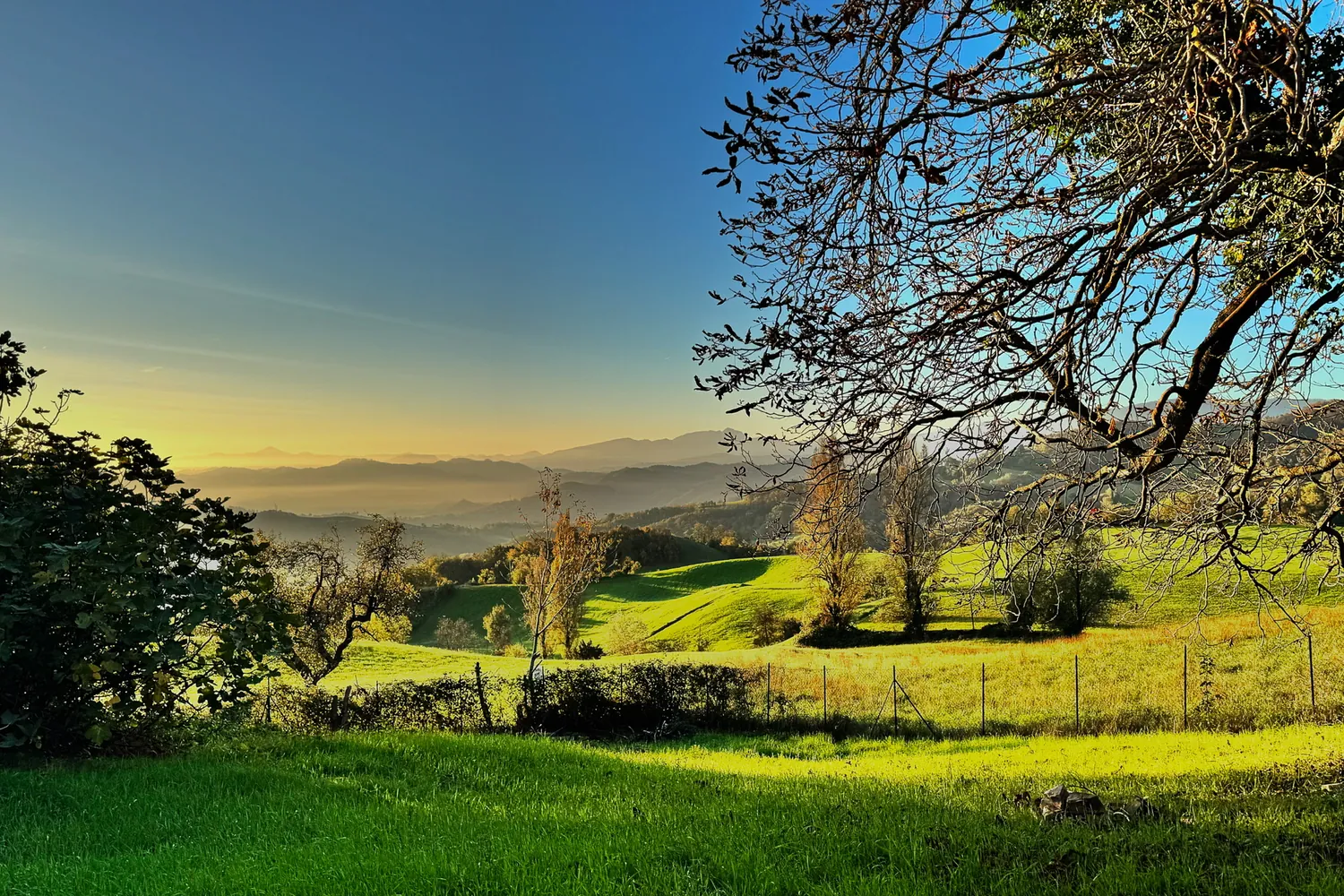 Tramonto dorato sulle colline boscose intorno a Urzano, con il cielo che sfuma dall'arancio all'azzurro sull'Appennino di Parma.